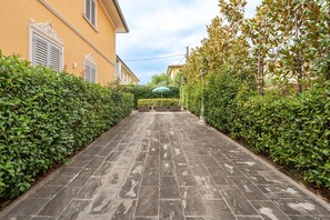 Un camino pavimentado bordeado por setos verdes y un edificio amarillo con contraventanas blancas.