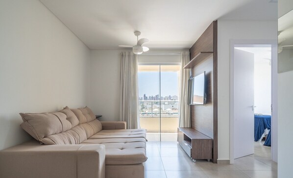A modern living room with a beige sectional sofa, a flat-screen TV mounted on a wooden wall, and a city view through large windows.