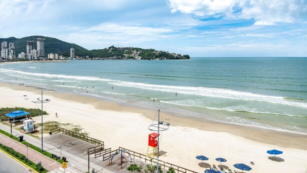 Une plage offrant une vue dégagée sur la mer, une route qui longe la plage, et une colline lointaine parsemée de bâtiments.