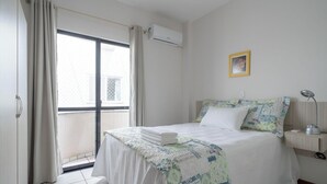 Une chambre à coucher avec un lit, des rideaux, un appareil de climatisation, une table de chevet avec une lampe et un tableau encadré au mur.