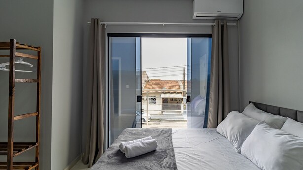 Ett hotellrum med en säng, en hylla i trä och utsikt över ett bostadsområde genom en glasdörr.