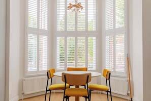 Un comedor con una mesa redonda y cuatro sillas amarillas, ventanas amplias con contraventanas blancas y suelo de madera.