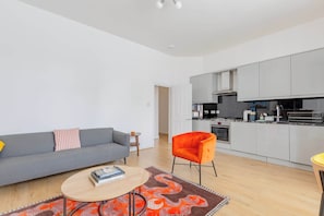 Un apartamento moderno con un sofá gris, una pequeña mesa redonda, una silla naranja llamativa, y una zona de cocina con armarios grises y una estufa negra.