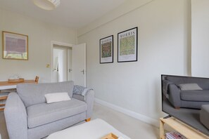 A living room with a grey sofa, a white armchair, a coffee table, and a TV on a wooden stand. There are two framed pictures on the wall and a door leading to another room.