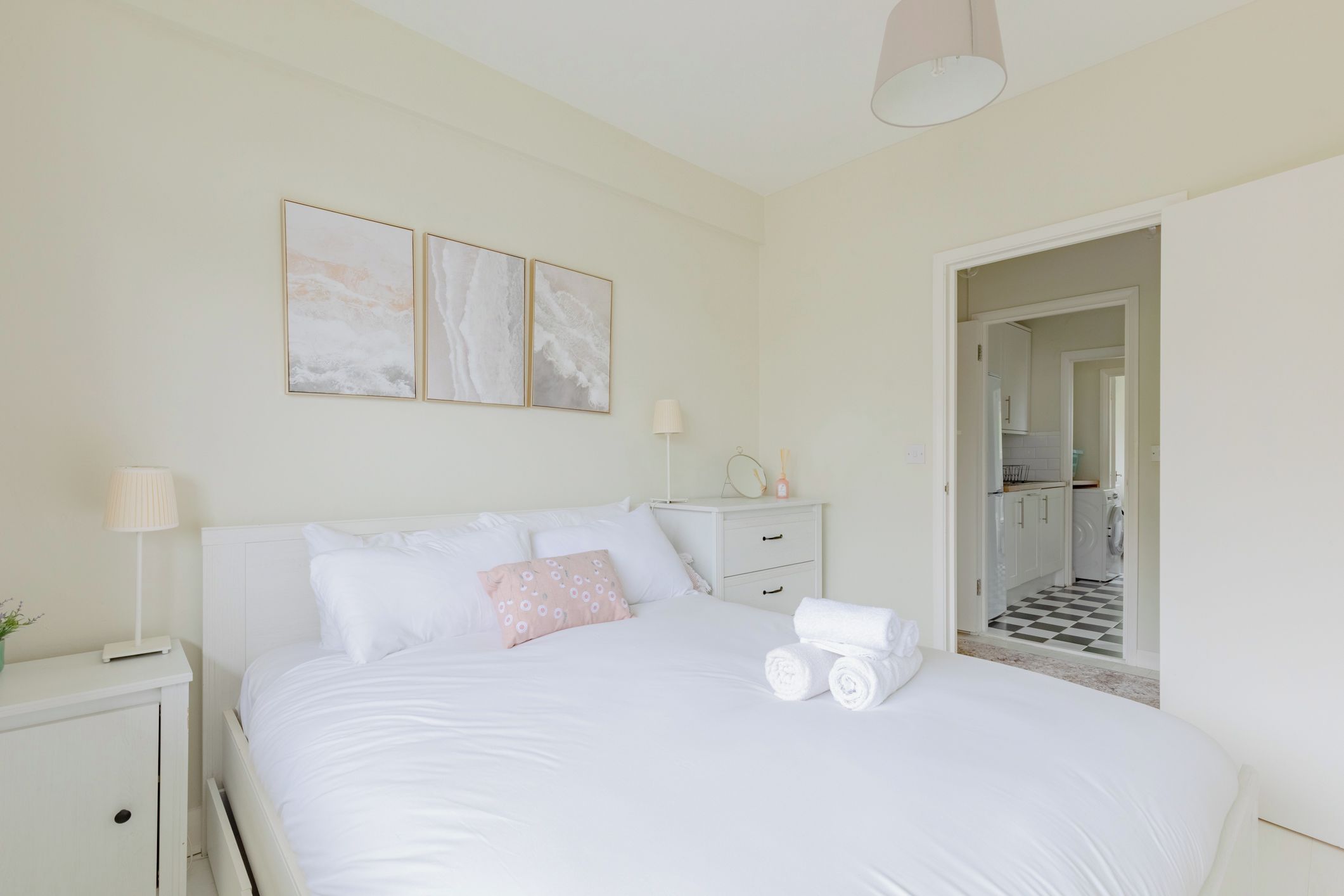 Une chambre à coucher comprenant un lit, une table de chevet, une lampe et une œuvre d’art. Une porte donne accès à un espace cuisine.