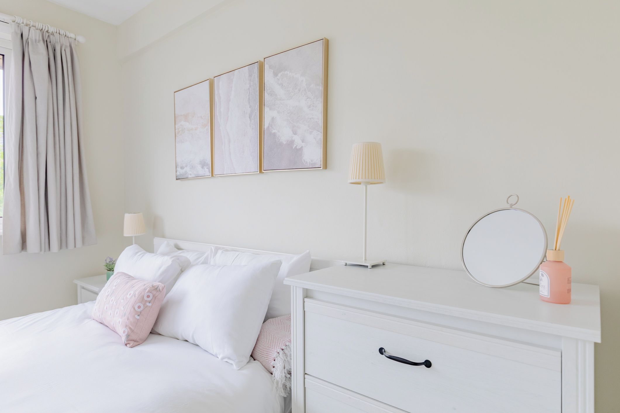 A neatly arranged bedroom with a bed, bedside table, and a dresser. There are three framed artworks on the wall above the bed.