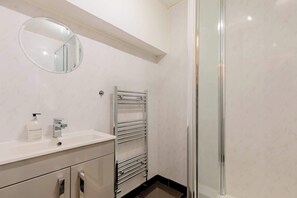 Casa de banho com toucador branco, espelho redondo, porta-toalhas e cabine de duche em vidro.