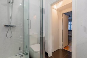 Un baño moderno con ducha acristalada, inodoro blanco y puerta visible que da a una zona de cocina.