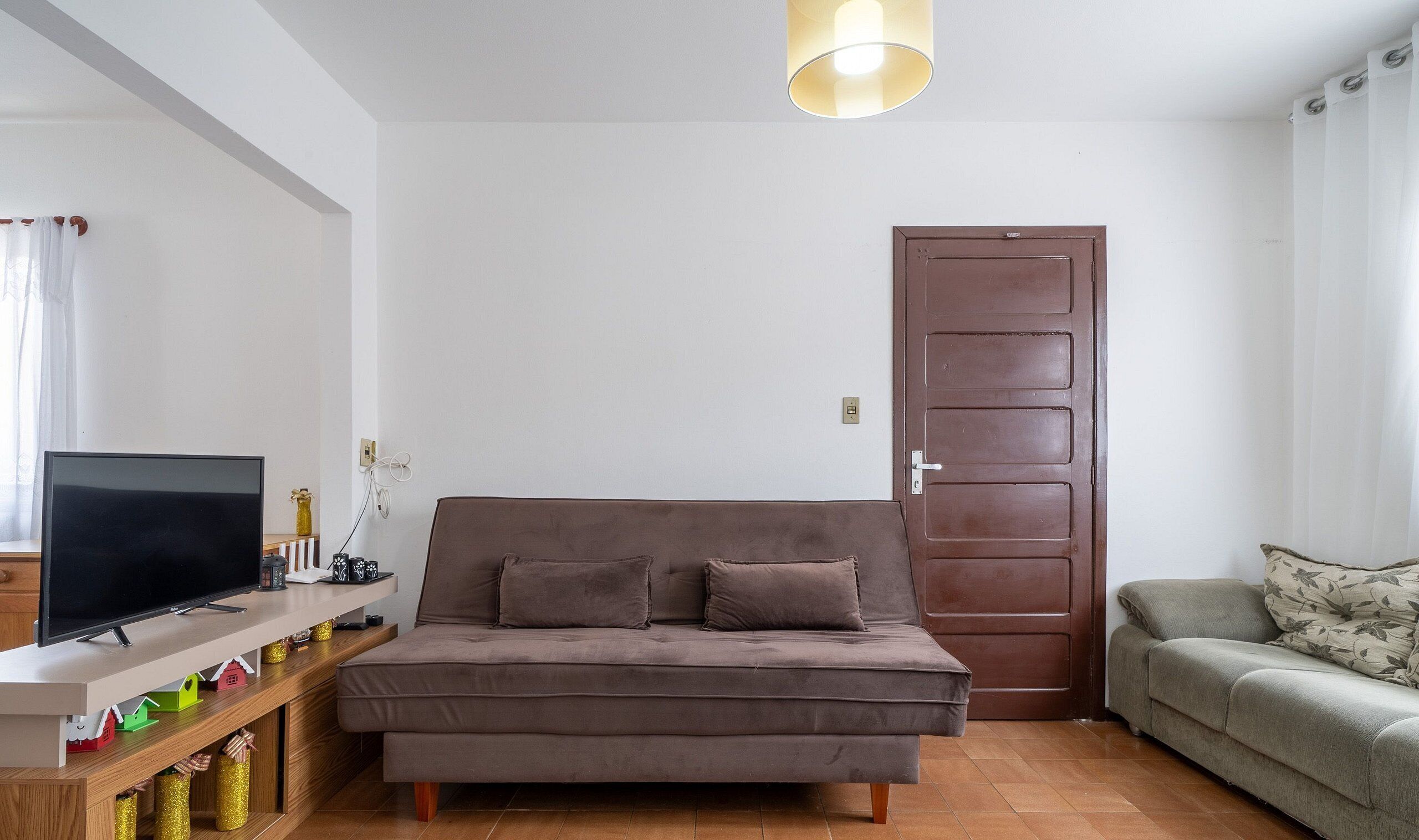 A living room with a brown sofa, a television on a stand, and a wooden shelf with decorative items.