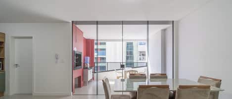 A modern living room with a dining area, a kitchen visible through a glass partition, and a view of the cityscape.