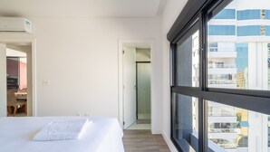 A modern hotel room with a large window, a bed, a desk, and a shower area. - Apto 2Q c/ churrasq. 500m da Meia Praia ROT1001 (Itapema)