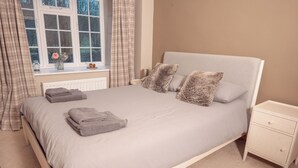 A neatly made bed with a grey duvet, two pillows, and folded towels on a white headboard. A small bedside table with a drawer is to the right.