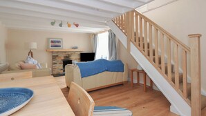 A cozy living room with a fireplace, a wooden staircase, and a dining area with a table and chairs. - 2 Porthilly Cottages (Wadebridge)