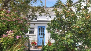 A quaint white building with a blue door, surrounded by lush greenery and flowering plants. - 2 Porthilly Cottages (Wadebridge)