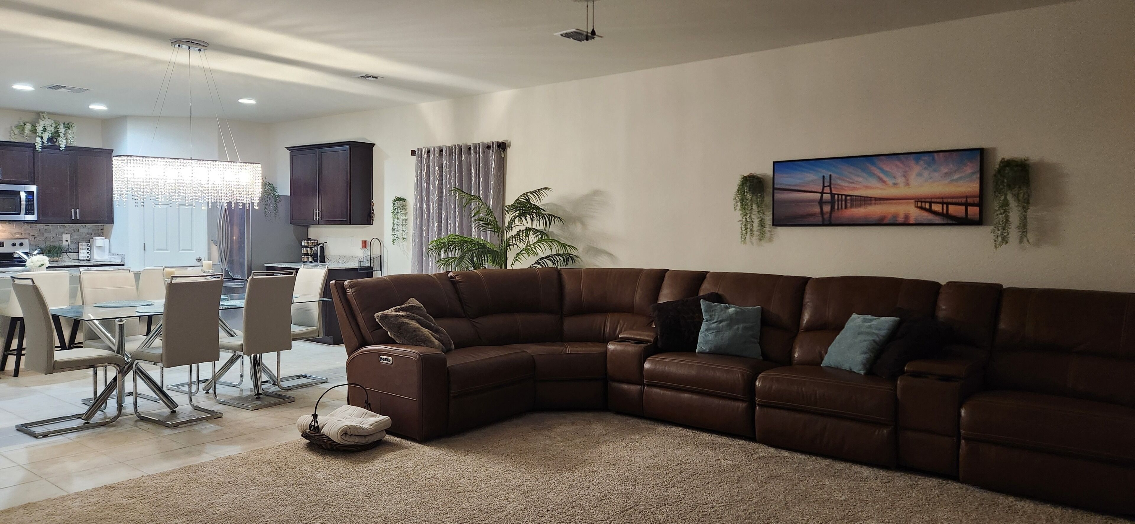 A modern living room with a brown sectional sofa, a glass dining table with chairs, a kitchen in the background, and a large window with curtains.