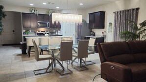 A modern kitchen with dark cabinets, a central island, and a dining area with a glass table and chairs. - Beautiful and Comfy 4-bedroom house with AC in Maricopa (Maricopa)