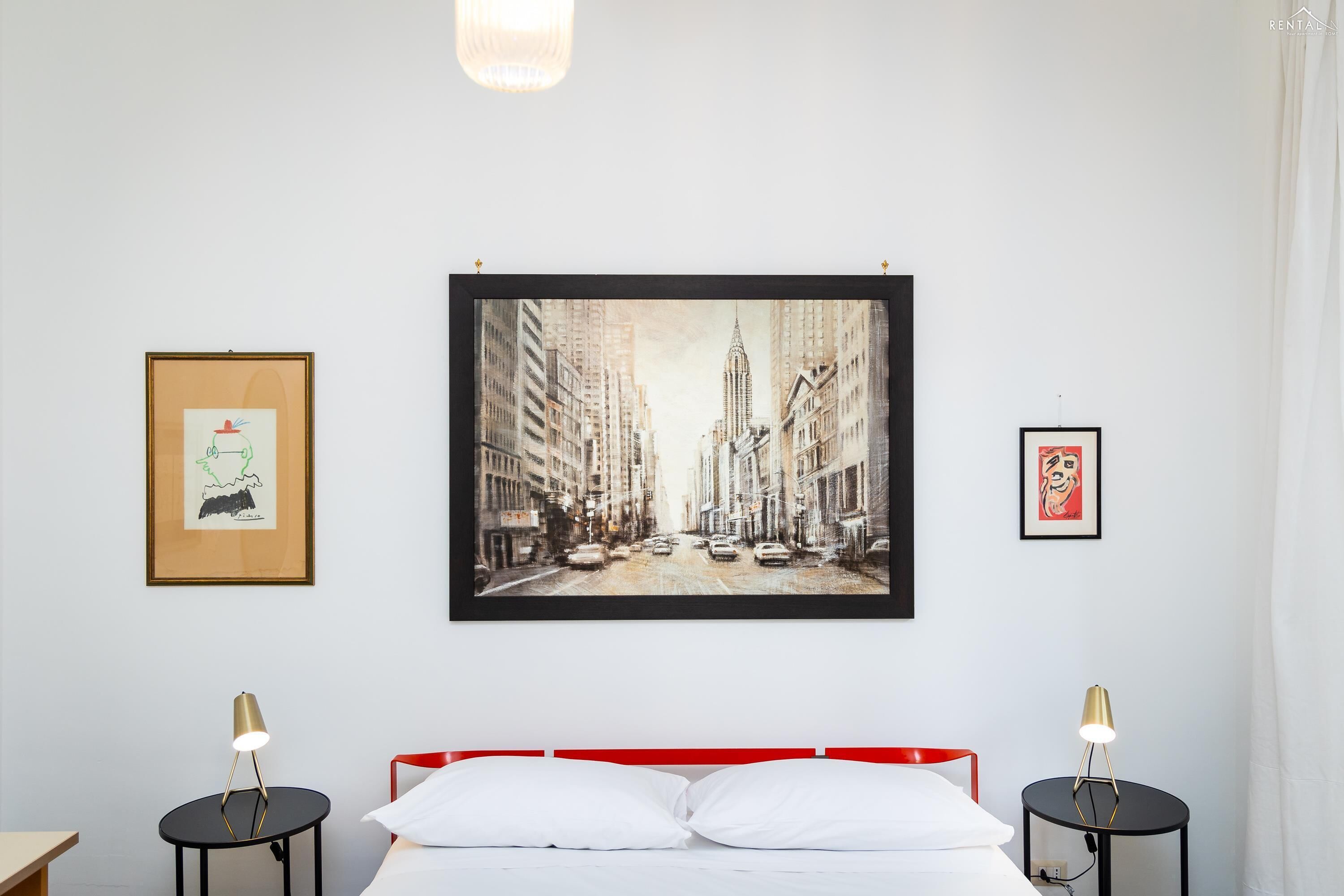Quarto de hotel com uma grande pintura emoldurada da paisagem urbana, uma cama com roupa de cama branca e duas mesas de cabeceira com luminárias.