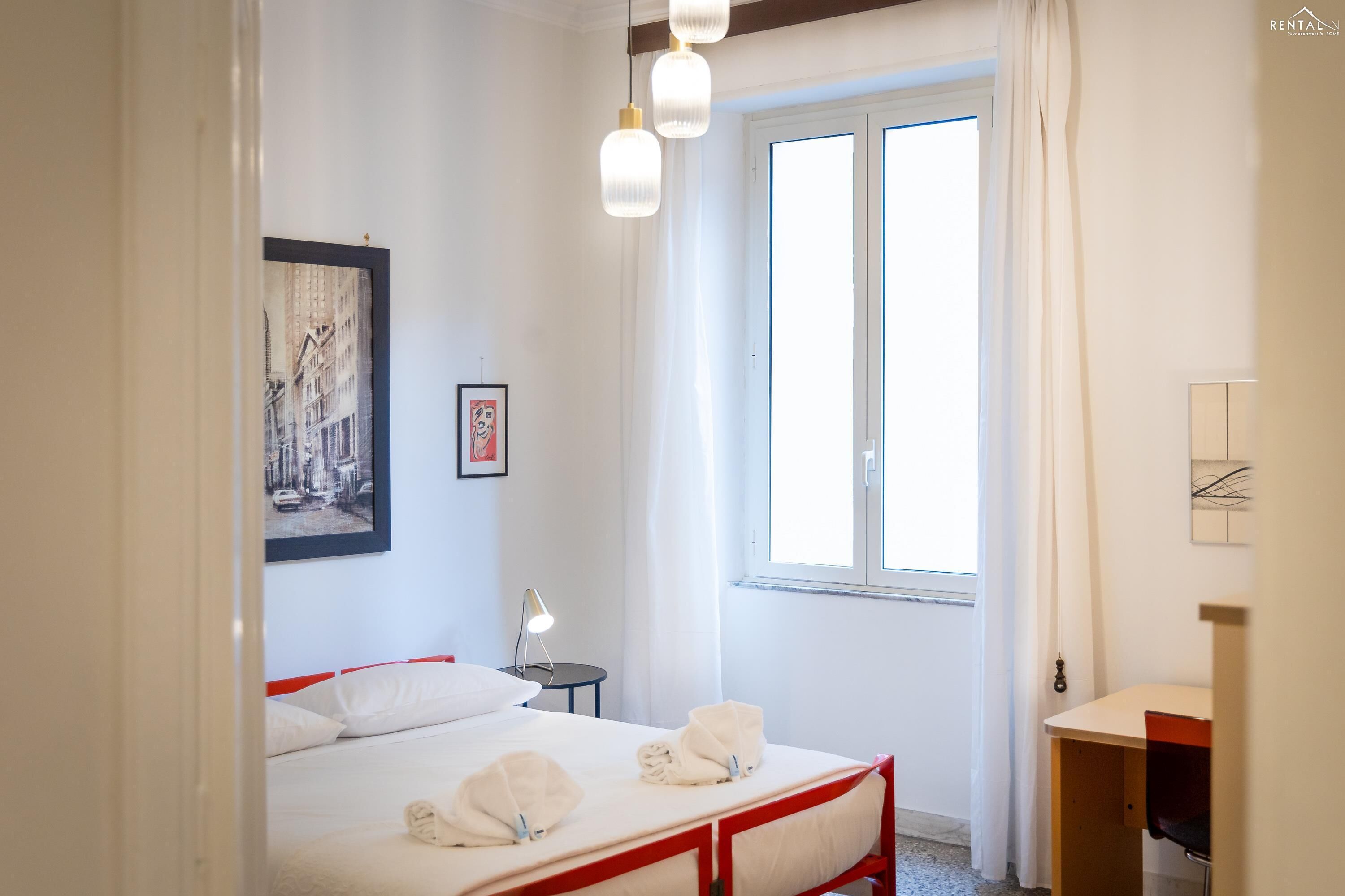 A hotel room with a bed, a bedside table with a lamp, a window with curtains, and a desk with a chair.
