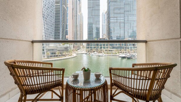Un balcon agrémenté de fauteuils en osier et d’une petite table, offrant une vue sur une marina où sont amarrés des bateaux et qui est bordée de bâtiments imposants.