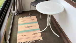 A small balcony with a white table and a gray chair, a woven rug, and a view of a building and trees.