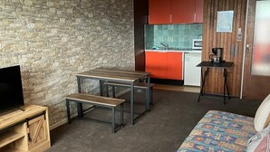 A compact living space with a stone wall, a wooden TV stand, a bench, a colorful sofa, a small kitchen with red cabinets, and a hanging light fixture.