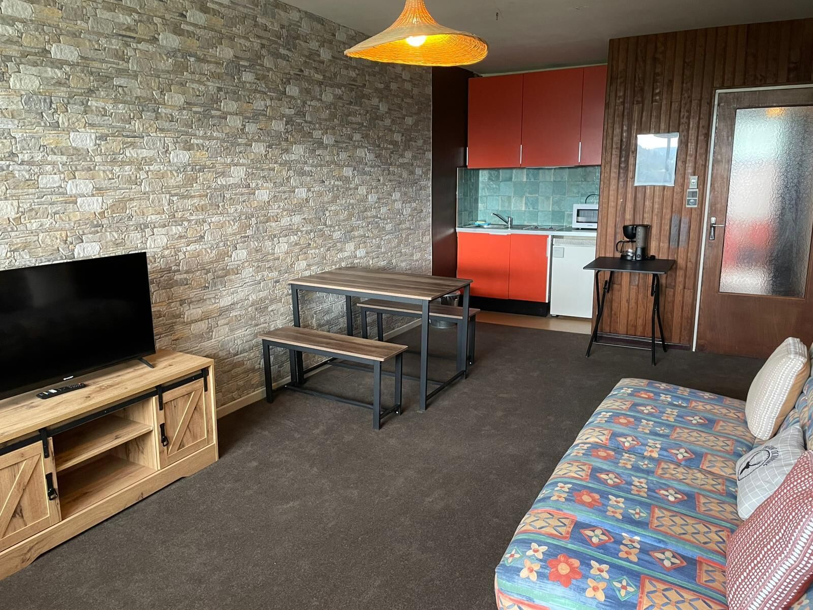 Un espacio habitable compacto con un televisor sobre un soporte de madera, una pequeña mesa de comedor con bancos, un sofá con cojines estampados, una zona de cocina con armarios rojos y una lámpara colgante.