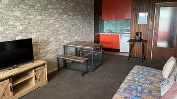 Un espacio habitable compacto con un televisor sobre un soporte de madera, una pequeña mesa de comedor con bancos, un sofá con cojines estampados, una zona de cocina con armarios rojos y una lámpara colgante.