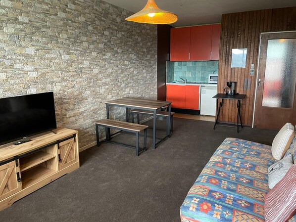 Un espacio habitable compacto con un televisor sobre un soporte de madera, una pequeña mesa de comedor con bancos, un sofá con cojines estampados, una zona de cocina con armarios rojos y una lámpara colgante.