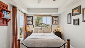 A bedroom with a bed, curtains, a ceiling fan, wall-mounted picture frames, and a window with a view of palm trees.
