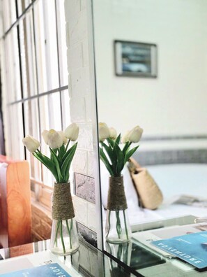 Une fenêtre ornée de tulipes blanches dans des vases en verre, un tableau encadré accroché au mur, et un aperçu d’une pièce où l’on aperçoit une chaise en bois et un petit livre bleu posé sur le rebord de la fenêtre.