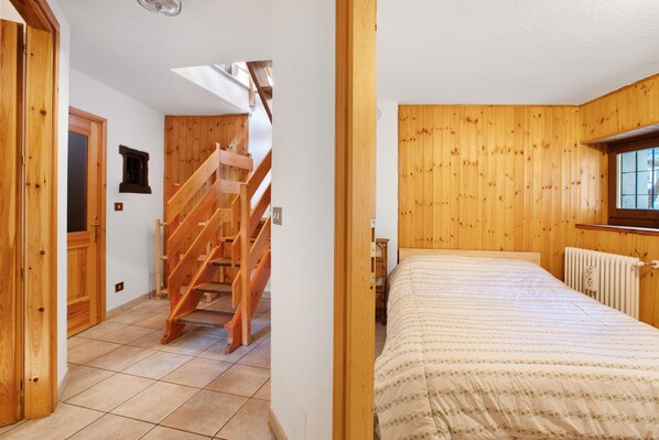 A bedroom with a wooden staircase, a bed with a patterned bedspread, and a window with blinds.