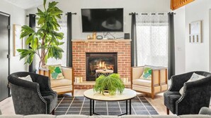Un salon moderne avec une cheminée en briques, deux fauteuils, un canapé, une table basse et une grande plante.