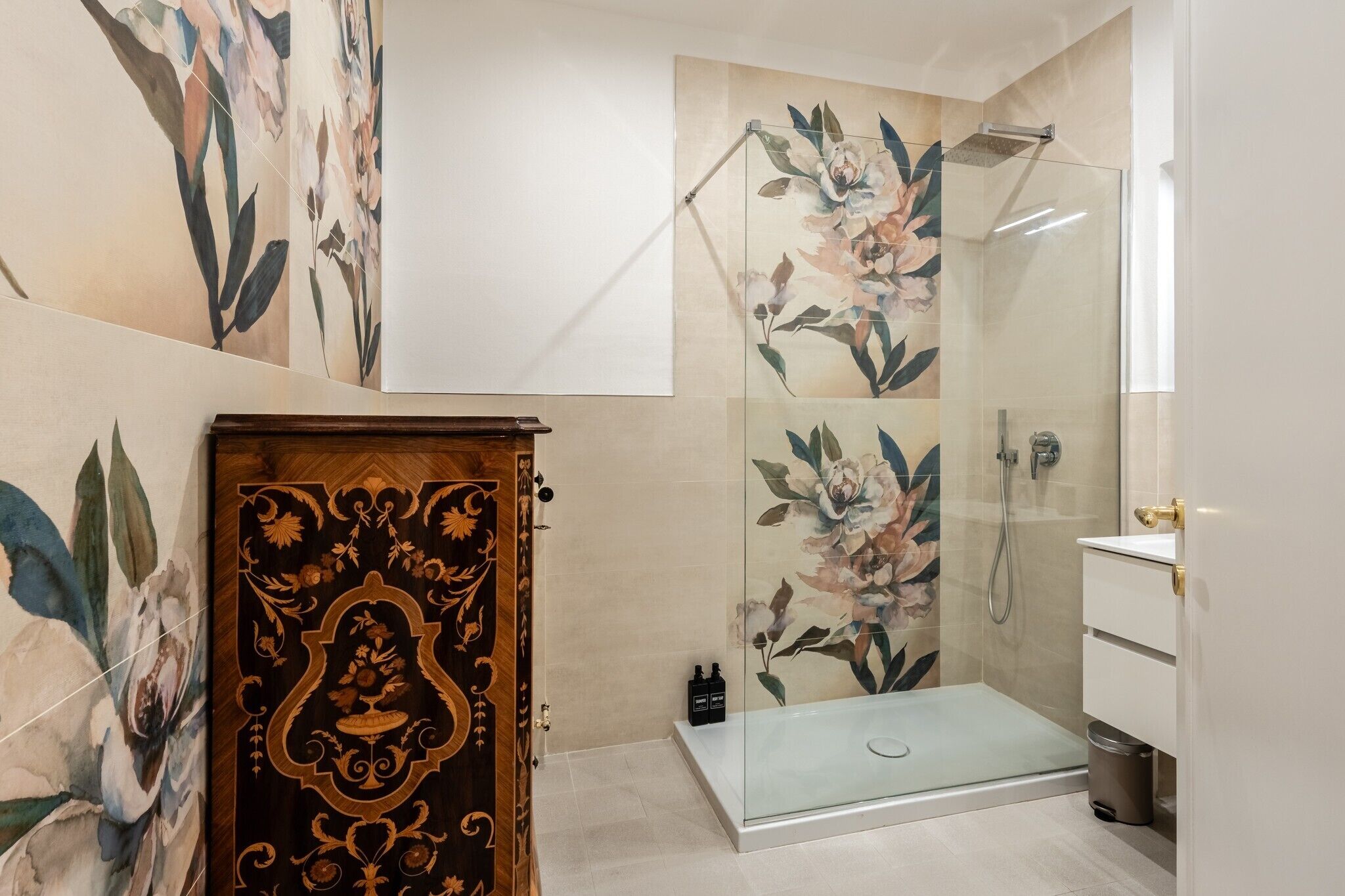 A modern bathroom with a glass shower, a wooden cabinet with intricate carvings, and floral wall art.