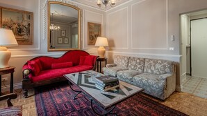Ein Zimmer mit einem roten Sofa, einem grau gemusterten Sofa, einem Marmortisch mit Büchern, einer Chandelüster und einem großen Spiegel.