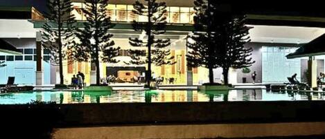 Un hotel con zona de piscina, mobiliario exterior y una entrada bien iluminada.