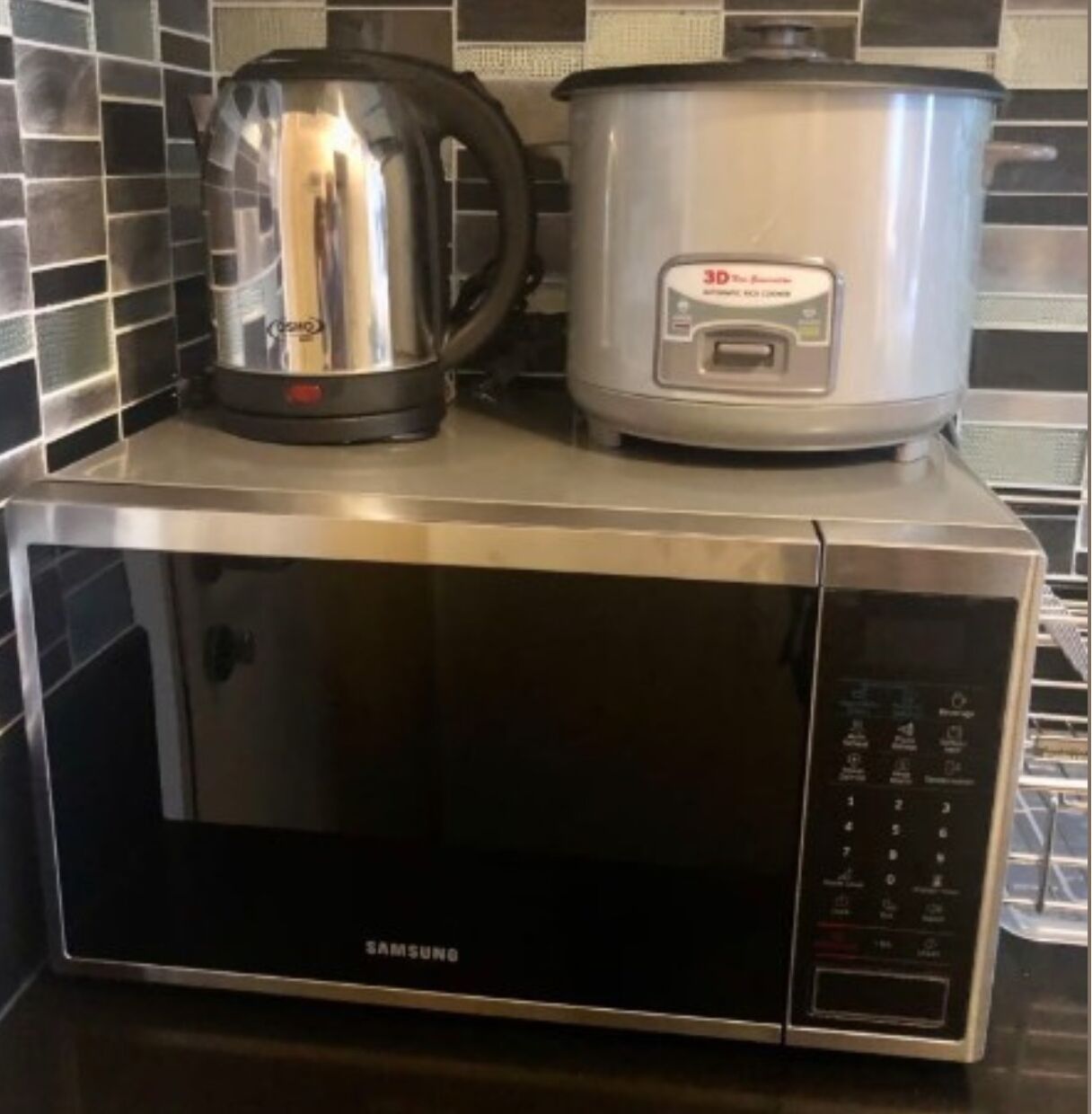 A Samsung microwave oven on a kitchen counter with a kettle and a slow cooker on top.
