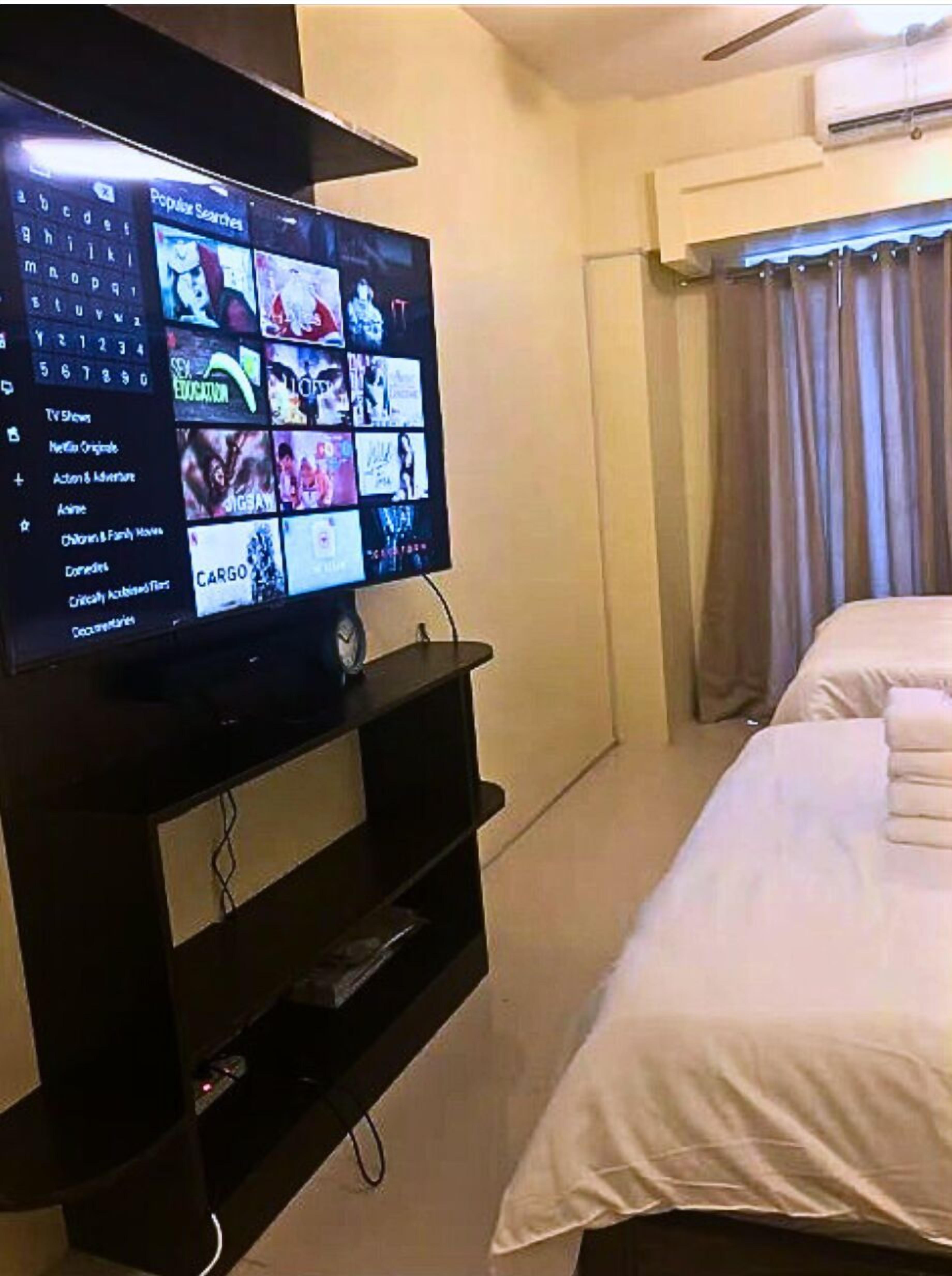 A hotel room with a TV displaying a movie selection menu, a bed, and an air conditioning unit.