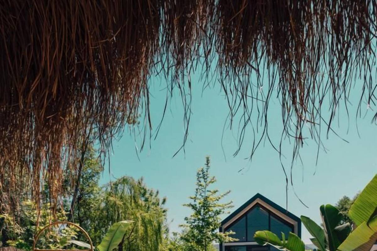 Una terraza de madera con piscina, una silla de descanso, una mesa con bebidas y una casa al fondo.