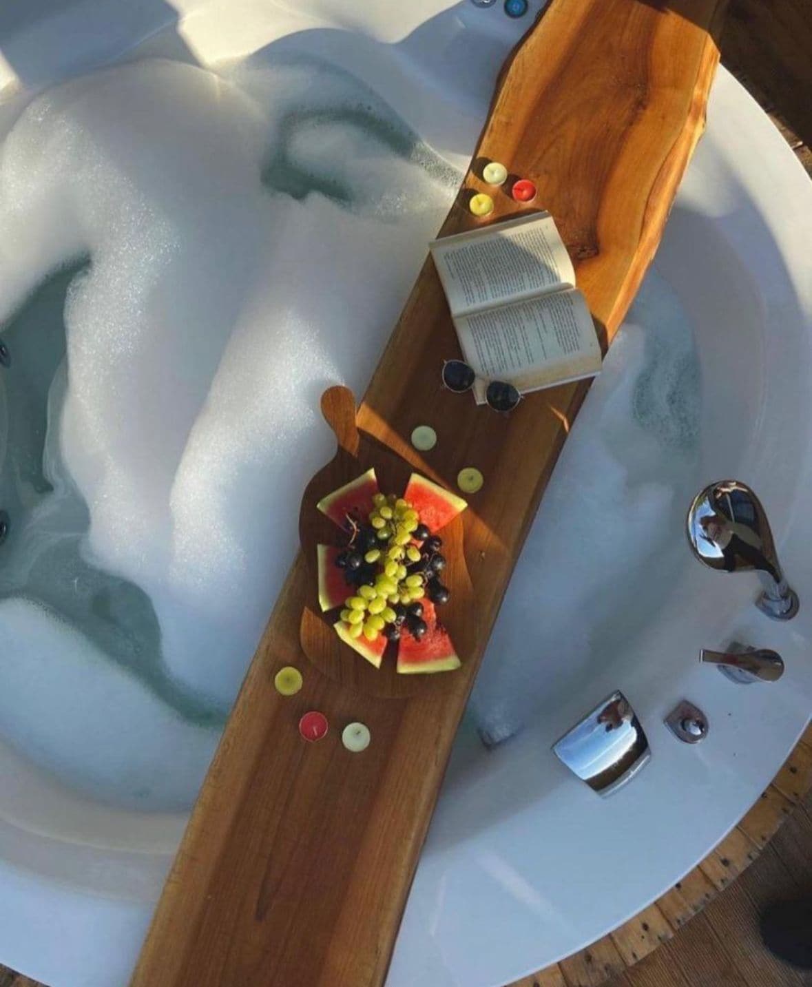 A hot tub with a wooden tray holding a book, a pair of sunglasses, and a bowl of fruit.