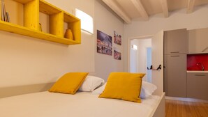 A bedroom with a bed, two pillows, a wooden shelf with books, and a red kitchen area.