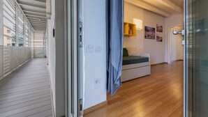A corridor with a sliding door leading to a room with a bed, a small table, and a wall-mounted shelf with items.