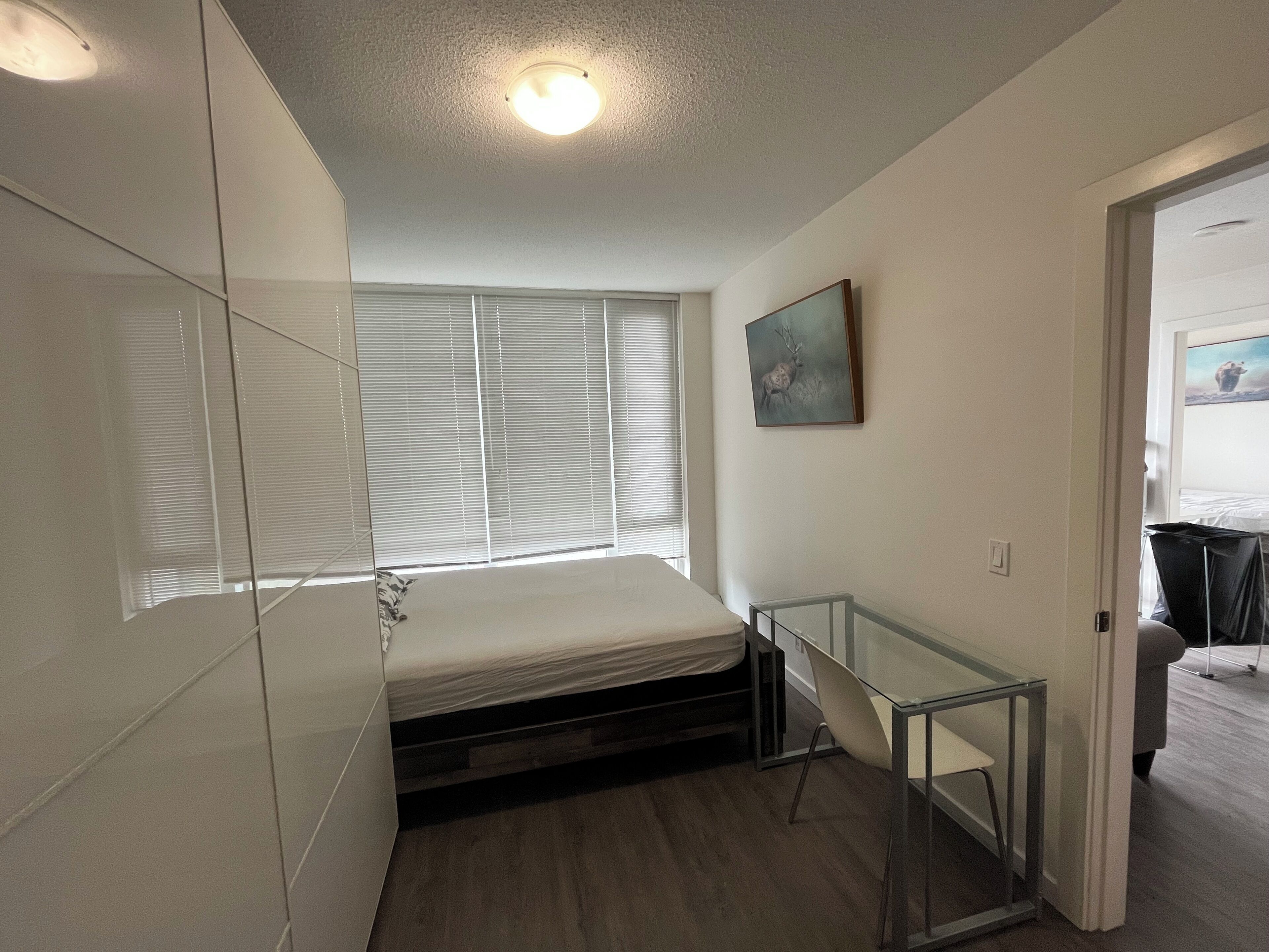 Een compacte slaapkamer met een bed, een glazen tafel en een raam met jaloezieën.