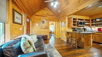 A cabin interior with a wooden ceiling, a living area with a couch and pillows, a kitchen with a microwave and bar stools, and a dining area with a table and chairs.