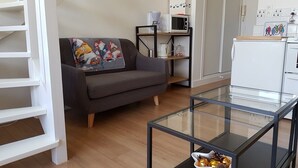 Una sala de estar moderna con un sofá gris, una mesa de centro de vidrio y una cocina con electrodomésticos blancos.