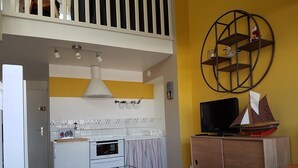 Cocina compacta con un refrigerador blanco, horno y una pequeña mesa de comedor con un tazón. Un gabinete de madera con un televisor y un modelo de barco está a la derecha.