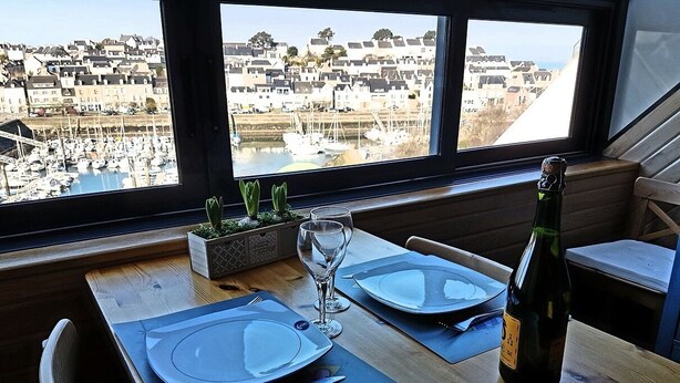 Mesa de comedor servida con platos, cubiertos y una botella de vino, con vistas a un puerto deportivo con barcos y edificios.