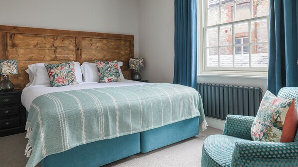 A bedroom with a large bed, a wooden headboard, a window with blue curtains, and a patterned armchair.