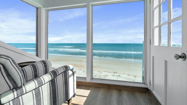 A room with a large window overlooking a beach, a striped sofa, and wooden flooring.