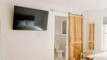 Un dormitorio con cama, televisor en la pared, armario de madera y baño visible a través de una puerta abierta.