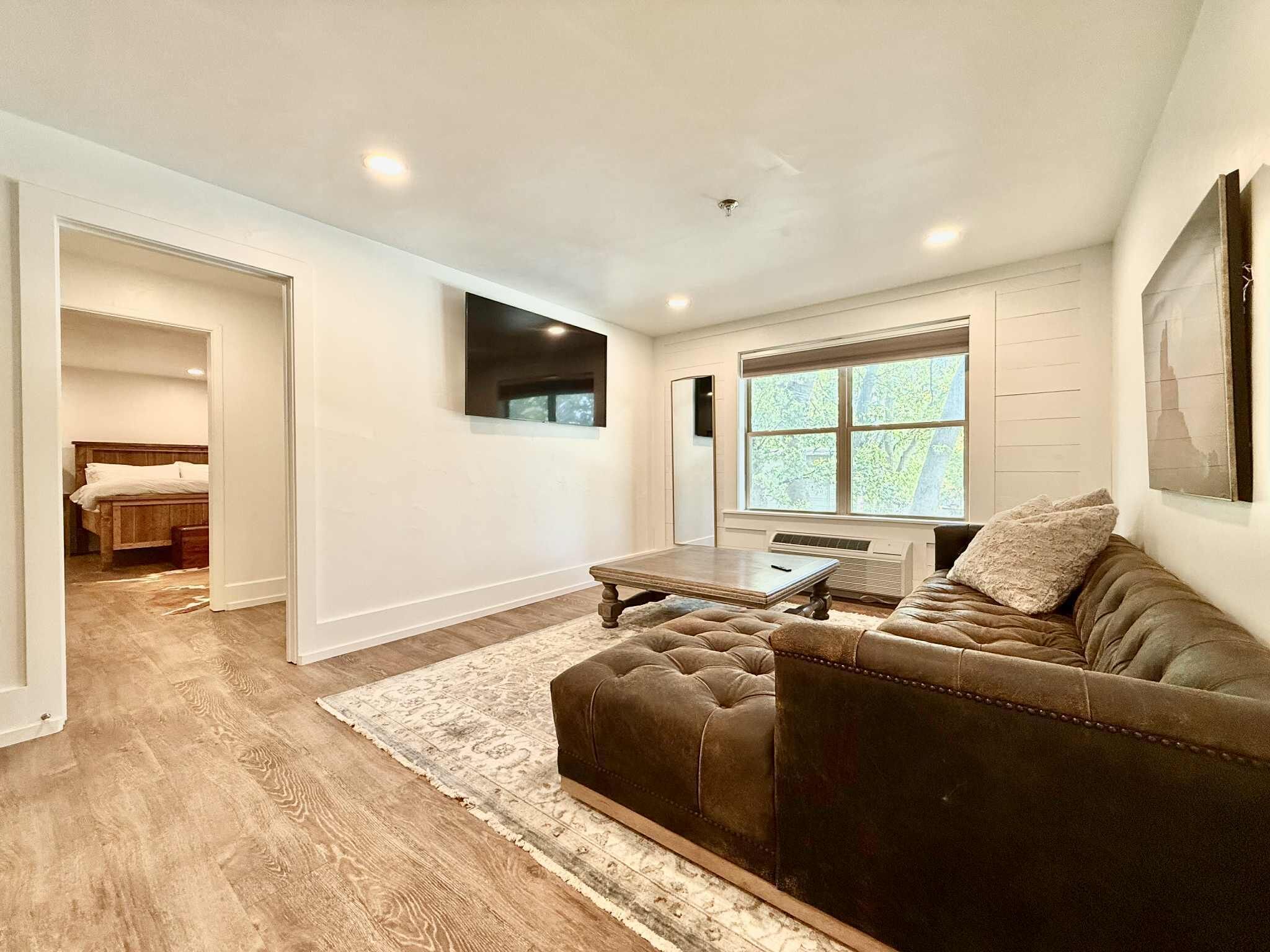 A living room with a brown leather sofa, a wooden coffee table, and a large window with a view of trees.